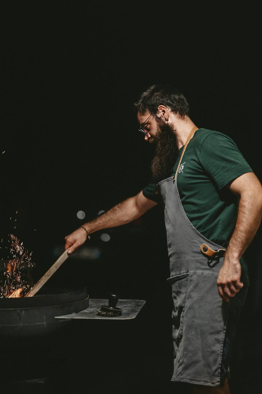A chef tending an open fire at Embers and Oak, sparks rising into the dark.